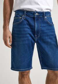 Pantalones cortos de mezclilla en azul oscuro con cinco bolsillos, trabillas para el cinturón y costuras en contraste. La tela tiene una textura suave y un ligero desgaste en la parte frontal.