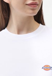 Hvid bomulds t-shirt med rund hals, der har et farverigt broderet logo på venstre brystområde. Glat tekstur.