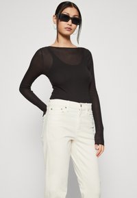 Top de manga larga negra acanalada con un escote amplio, combinado con jeans de mezclilla blancos de tiro alto. La modelo lleva unas gafas de sol negras angulares y llamativas.