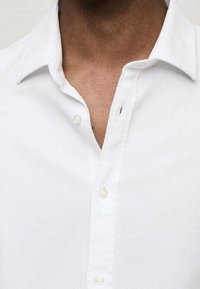 Camisa blanca de cuello con una tela texturizada, cierre de un solo botón y una pequeña apertura cerca del área del cuello. Detalle de diseño simple.