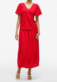 Czerwony dwuczęściowy zestaw z topem o dekolcie w serek i krótkimi bufkami oraz dopasowaną długą spódnicą. Materiał wydaje się gładki i lekki.