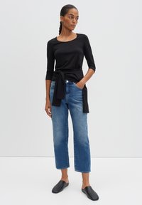 Zwarte lange mouwen top met geknoopte detail, lichtblauwe rechte jeans en zwarte instappers tegen een neutrale achtergrond.