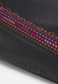 Sac à main en cuir noir avec des détails brodés colorés : fils roses, violets et oranges dans des motifs complexes sur le côté. Surface texturée.