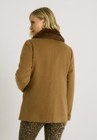 Abrigo de mezcla de lana marrón con cuello de piel sintética, ajuste entallado y mangas largas, combinado con pantalones de estampado de leopardo negro y marrón.