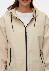 Sieviete valkā bēša krāsas kapuci ar melnu rāvējslēdzēju un aukliņām virs baltas krekla, redzama no pleciem līdz viduklim.