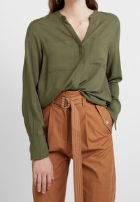Camicia a maniche lunghe verde oliva con scollo a V, due tasche sul petto, abbinata a pantaloni marroni a vita alta con cintura e tasche laterali.