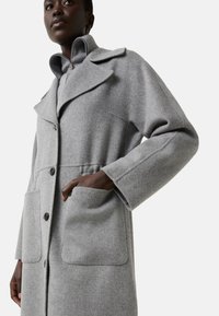 Kvinde iført en lys grå uldfrakke med knapper, bred krave og store forlommer, som poserer med den ene hånd i en lomme.