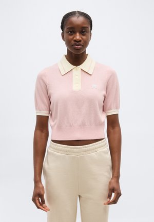 Vrouw met gevlochten haar, gekleed in een aansluitende roze polo met korte mouwen en beige broek met elastische tailleband, tegen een effen achtergrond.