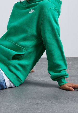 Person iført en grøn Nike hættetrøje med en frontlomme, siddende på en grå gulv med den ene arm strakt bagud for støtte.