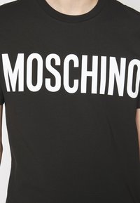 T-shirt en coton noir avec un grand texte blanc "MOSCHINO" sur la poitrine, design classique à col rond, texture lisse.