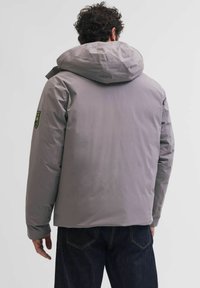 Šedá zateplená bunda s kapucí, s jemnou texturou a logem na levém rameni. Jednoduchý design, délka po kolena a dlouhé rukávy.
