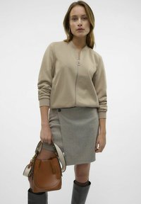 Sweatshirt beige à fermeture éclair, jupe grise en chevrons, sac seau marron avec une bandoulière crème, et bottes noires à hauteur de genou. Design minimaliste avec des détails subtils.