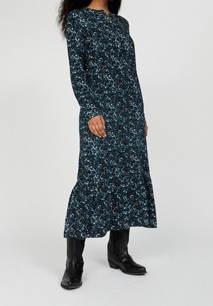 Frau trägt ein langärmeliges, dunkelblumiges Midikleid kombiniert mit schwarzen Lederstiefeln bis zur Mitte der Wade, stehend vor neutralem Hintergrund.