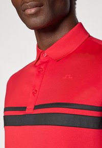 Červená polo košeľa so štruktúrovanou tkaninou, tradičným golierom a čiernymi pruhmi cez hruď, s piatimi gombíkmi na zapínanie.