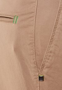 Béžová látka s hladkou texturou, zdobená zeleným akcentním prošíváním a malou značkovou visačkou. Design zahrnuje plochou kapsu a detaily švů.