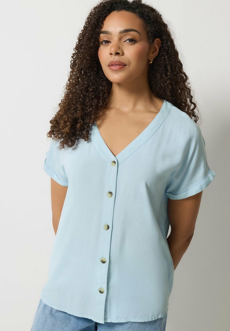 Femme aux cheveux bouclés portant un chemisier bleu clair à manches courtes et boutonné, posant devant un fond clair uni.