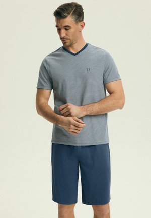 Muž v námorníckom a bielom pruhovanom tričku s krátkym rukávom a výstrihom do V a tmavomodrých šortkách si upravuje ľavý zápästie.