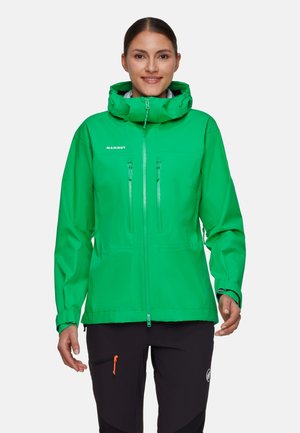 Kvinde iført en knaldgrøn hættejakke til udendørs brug med lynlåse på brystlommerne og sorte bukser med orange lynlåstræk.