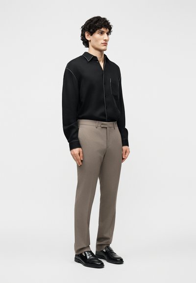 Modèle masculin aux cheveux bouclés portant une chemise noire à col, un pantalon beige sur mesure et des mocassins en cuir noir, debout devant un fond uni.