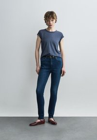 Fiatal személy egyszínű fal előtt áll, rövid ujjú szürke pólót, magas derekú sötétkék szűk farmert és barna lapos cipőt viselve.