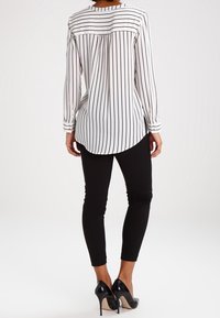 Blouse à manches longues rayée noire et blanche, avec un ourlet arrondi et un pli au dos, associée à un pantalon noir ajusté et des escarpins noirs.