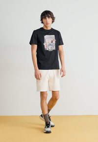 Black cotton t-shirt with a colorful graphic print, paired with light cream shorts. Model wears gray socks and black athletic shoes.