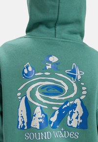 Grön hoodie med blå och vit grafik av stenar, spiralvågor, musiknot, ögonfigur, geometrisk form och texten "Sound Waves" på ryggen.