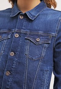 Mörkblå denimjacka med spetsig krage, kopparfärgade knappar och två bröstfickor med klaffdetalj. Rak passform med sömnadsdetaljer.