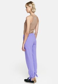 Brązowy bezrękawnik i fioletowe luźne spodnie zawiązywane na kostkach, model nosi czarne klapki.