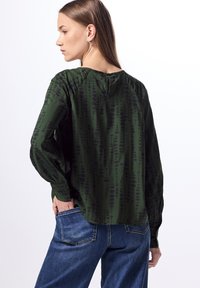 Blusa verde con un estampado sutil, mangas largas y un detalle de escote en forma de lágrima en la parte posterior, combinada con jeans de mezclilla azules. Textura de tela suave.