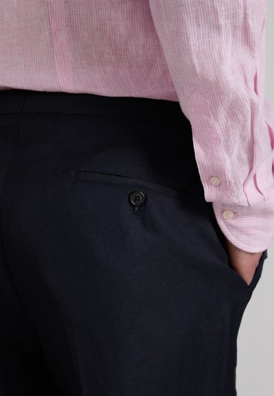 Man wearing dark navy trousers and a light pink, long-sleeve, buttoned shirt with hand in pocket, shown from waist to mid-thigh.