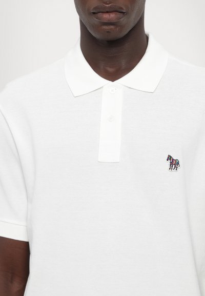 Valge polo särk tekstuuriga kangast, millel on kolmnupuline kinnituse ja väike mitmevärviline sebra embleem vasakul rinnal.