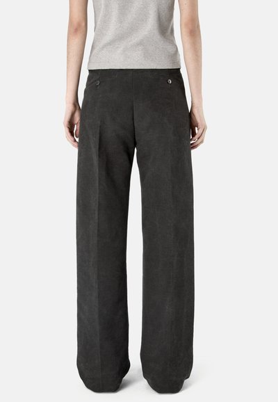 Kobieta ubrana w luźne, czarne spodnie z wysokim stanem i kieszeniami z tyłu zapinanymi na guziki, stojąca tyłem na jednolitym tle.