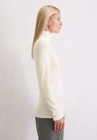 Crèmekleurige turtleneck-top van ribstof, nauwsluitend ontwerp, lange mouwen, gecombineerd met grijs gestreepte broek, vlakke lichte achtergrond.