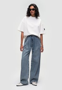 Camiseta blanca de corte oversize, combinada con jeans de pierna ancha en azul claro. La modelo lleva gafas de sol negras y zapatos blancos de punta con detalles en negro.