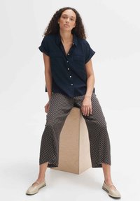 Marineblauwe overhemd met korte mouwen, met een patroon van brede broek met geometrische ontwerpen en beige platte schoenen, zittend op een houten kubus.