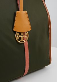 Sac vert foncé avec garniture en cuir rose et beige et un pendentif logo rond en métal doré fixé près du haut au centre.