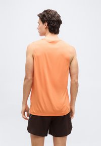 Νεαρός άνδρας με κοντά καστανά μαλλιά που φοράει πορτοκαλί αμάνικο αθλητικό μπλουζάκι και μαύρο σορτς, με την πλάτη προς το μέρος μας, σε απλό λευκό φόντο.