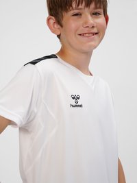 Lächelnder Junge mit weißem Hummel-Sportshirt, schwarzem Bienenlogo und schwarzen Chevron-Streifen auf der Schulter vor einfachem Hintergrund.