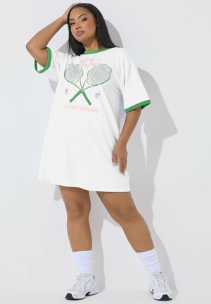 Mujer con vestido de tenis blanco y verde con un gráfico de raquetas cruzadas y el texto "Always Serving", posando con una mano en la cabeza.