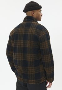 Uomo che indossa una giacca di pile a scacchi marrone e nera e un berretto di maglia nero, con il viso di lato e le mani nelle tasche, su uno sfondo bianco.