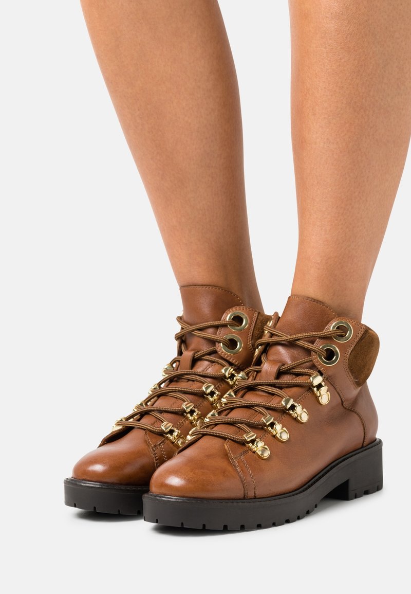 Bottines en cuir marron avec un bout rond, des détails en métal doré, des lacets texturés, et une semelle noire épaisse.