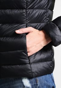 Černá prošívaná bunda s vodorovným prošíváním a kapsou na zip. Povrch je hladký a rukávy jsou těsné u zápěstí.