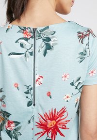 Světle modrý květinový top s zipem na zadní straně, s červenými a růžovými květinami na měkké strukturované tkanině. Krátké rukávy.