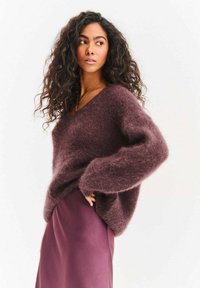 Bordowy, oversize sweter z dekoltem w serek, wykonany z puszystego materiału, zestawiony z gładką, lawendową spódnicą. Teksturowane, obszerne rękawy.
