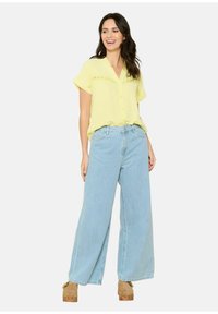 Camicia gialla chiara a maniche corte con chiusura a bottoni e dettagli in pizzo, abbinata a jeans blu chiaro a gamba larga e scarpe con platform color sabbia.