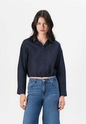 Chemise courte boutonnée bleu marine à manches longues, comportant des accents décoratifs sur le col. Associée à un jean en denim bleu.