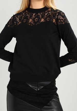 Czarna bluzka z długim rękawem z kwiatową koronkową wstawką na karczku i na ramionach, noszona z czarną skórzaną spódnicą z nakładką zamszową w kształcie litery V.
