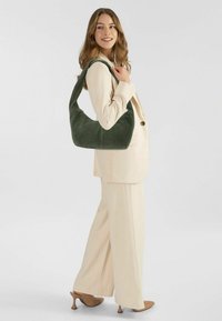 Borsa a spalla verde in suede con una forma curvilinea e un'ampia tracolla, abbinata a un completo crema. Texture liscia e dettagli hardware minimali.