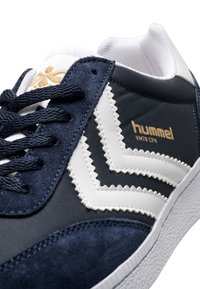 Marinblå sneakers med vita accenter och zigzag-mönster; tillverkade av mocka och textilmaterial; har präglad branding i guld.